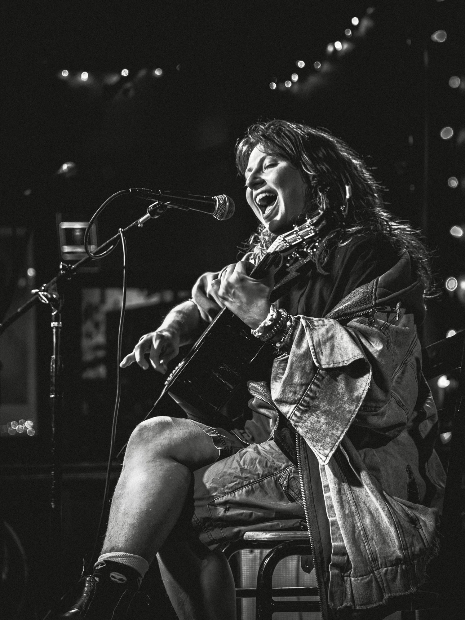 Black and white photo of a female musician passionately singing and playing guitar on stage at a live venue.