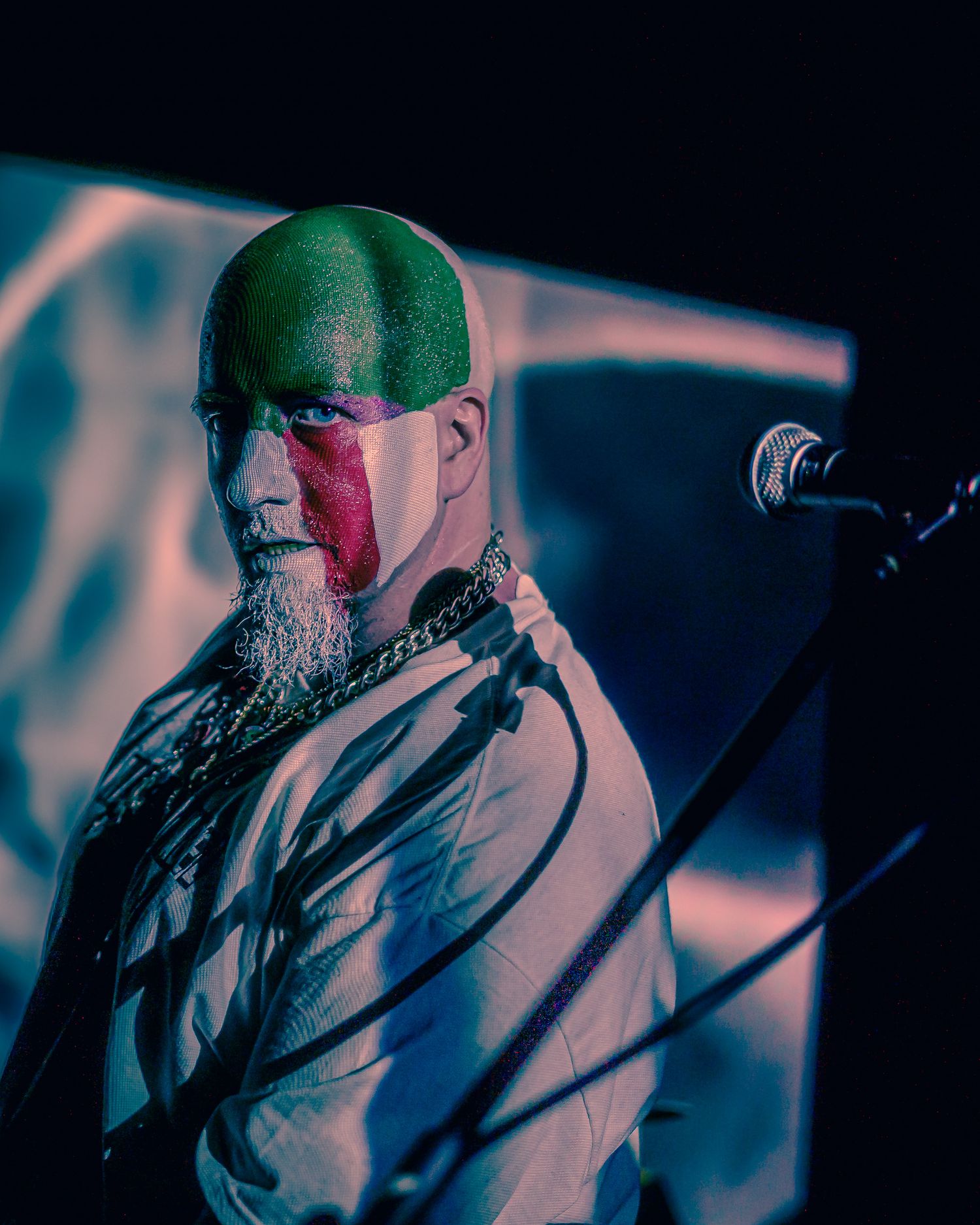 Performer with face paint and beard illuminated by dramatic lighting on stage with microphone stand in background.
