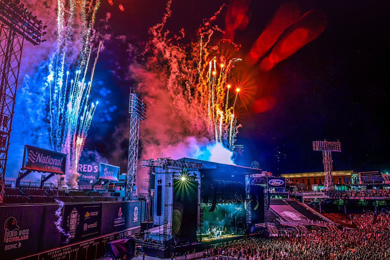 Spectacular fireworks and light show illuminate outdoor concert stage with large crowd during nighttime performance.
