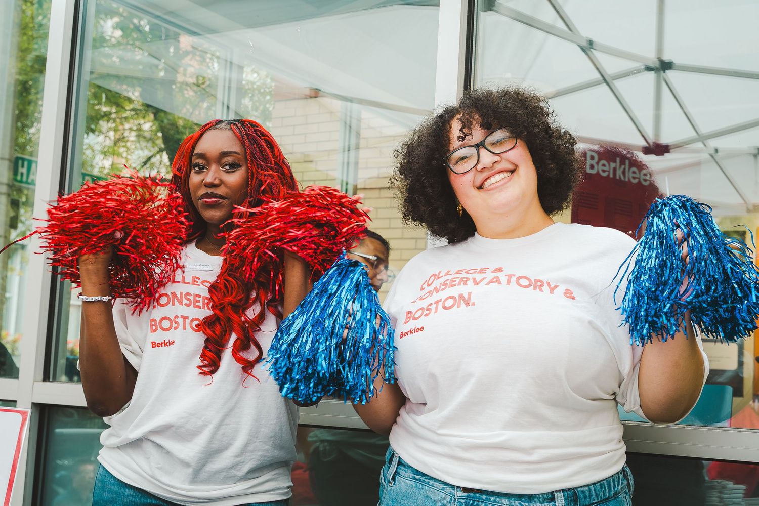 Two cheerful women in white Berkeley shirts hold red and blue pom-poms outdoors at a campus event.