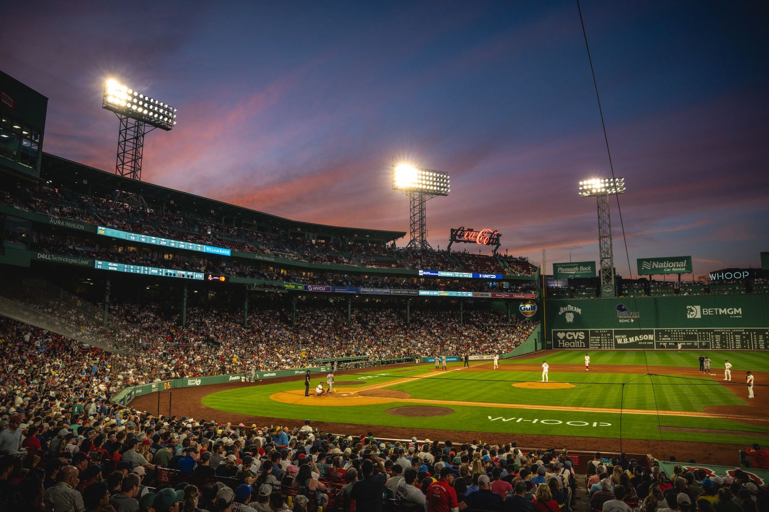 Fenway Park baseball stadium glowing under bright lights at twilight with a packed crowd and vibrant sunset sky.