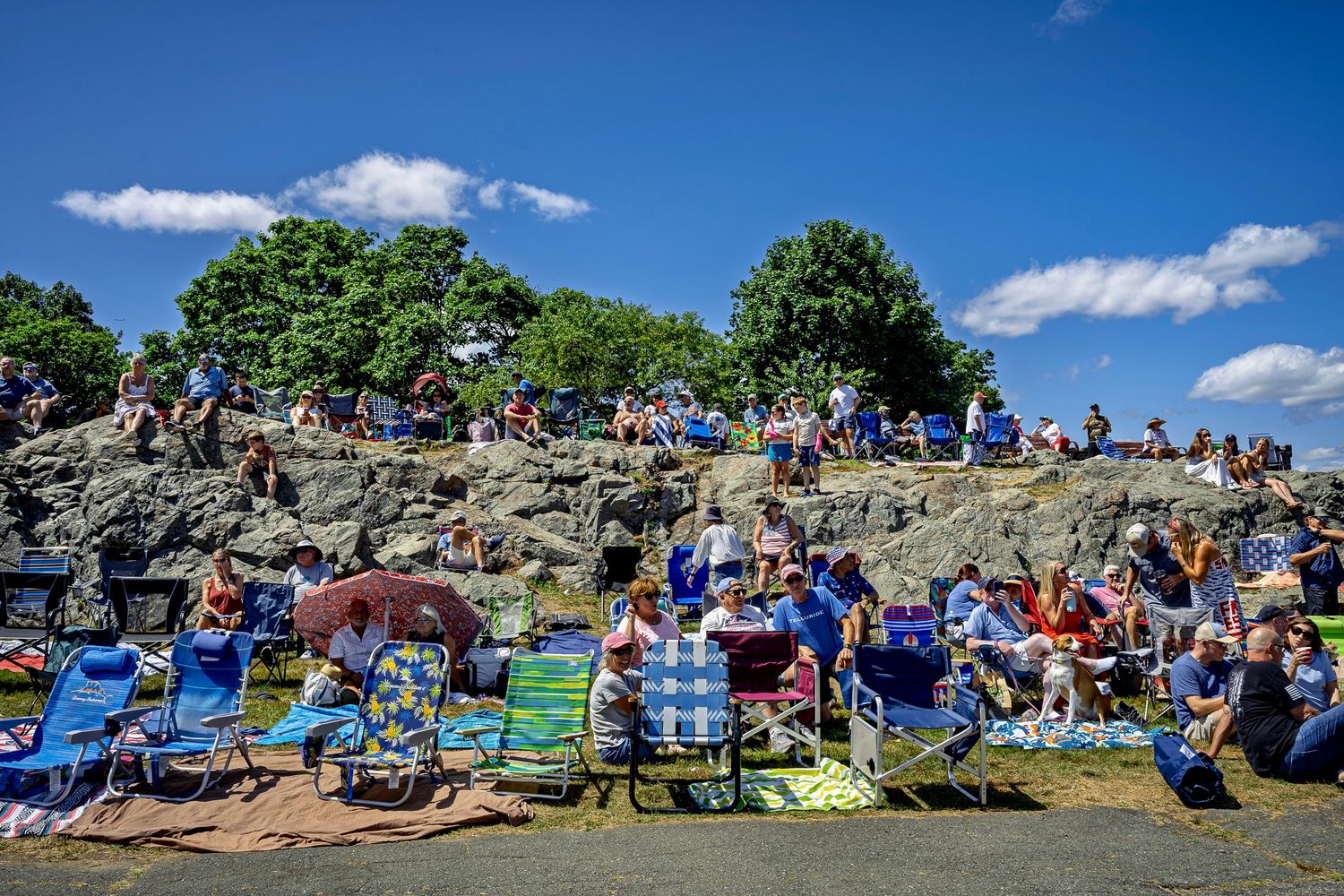 Crowd of people relaxing in lawn chairs on a rocky hillside on a sunny summer day with blue skies and white clouds.