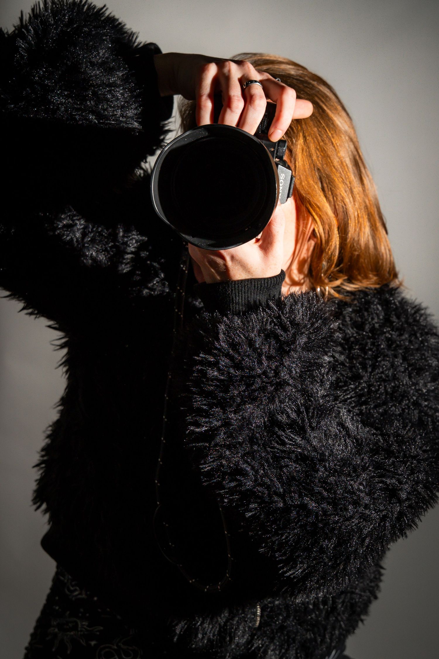 Red-haired photographer in stylish, fluffy black coat holding a mirrorless Sony camera up to their face against a gray background.