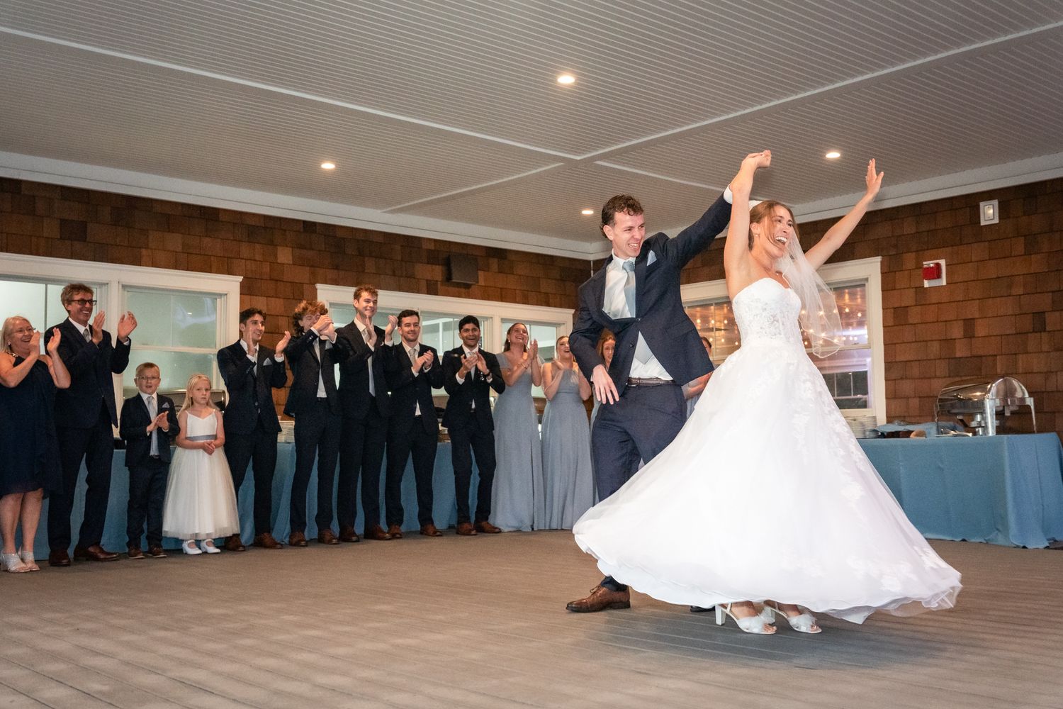 Wedding couple performs their first dance as newlyweds on the dance floor surrounded by guests at a reception.