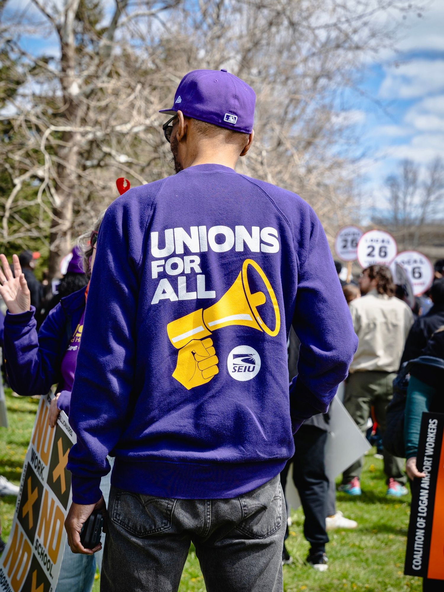 Person wearing purple SEIU Unions For All sweatshirt at labor rally with protesters holding signs outdoors.