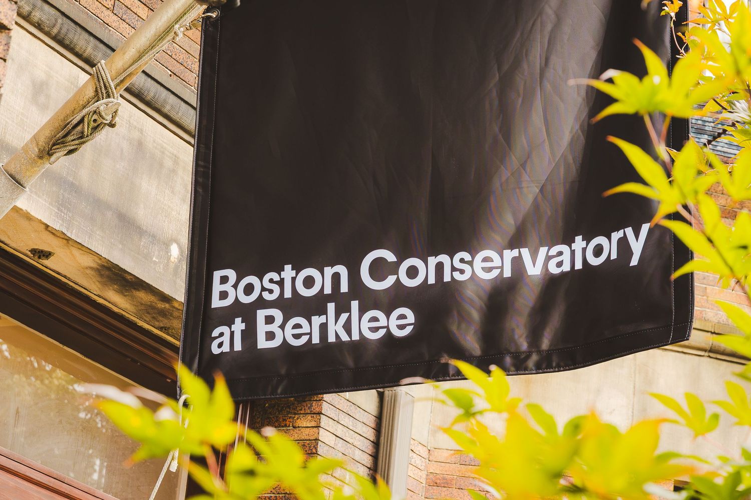 Black banner sign for Boston Conservatory at Berklee mounted on brick building exterior with green foliage.