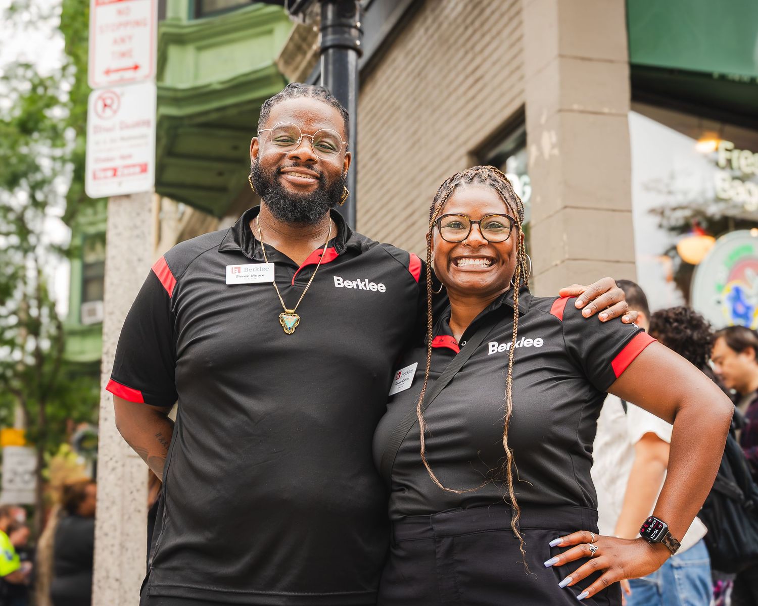 Two smiling employees in black Berklee polo shirts pose together outdoors at a street event.