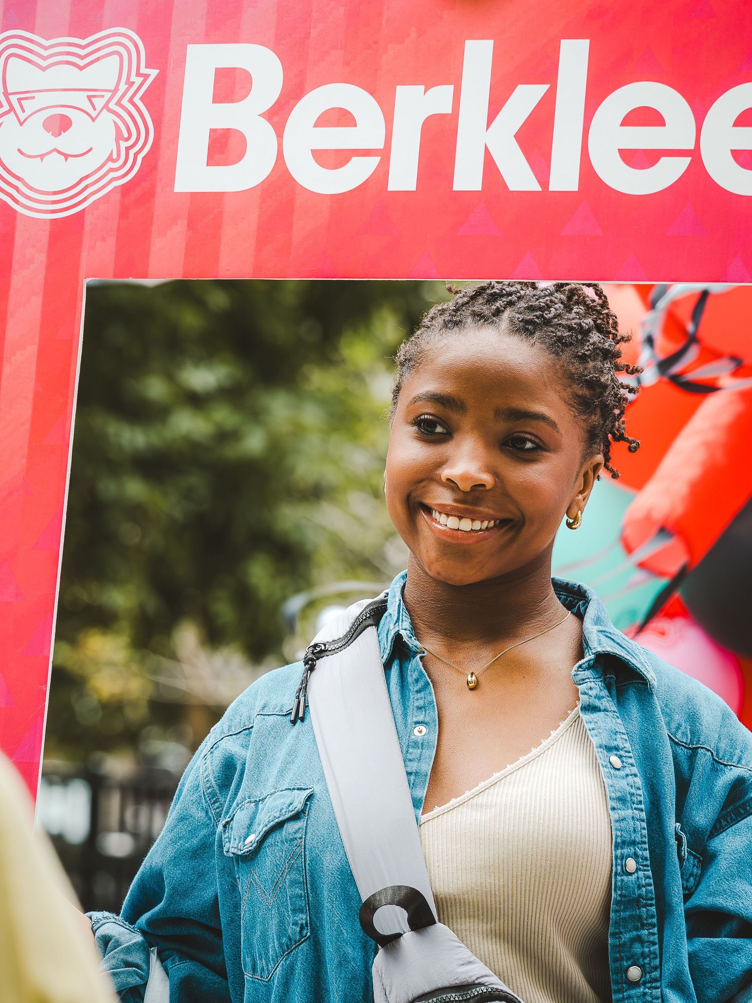 Smiling young woman with braids wearing a denim jacket near a Berklee College of Music banner outdoors.