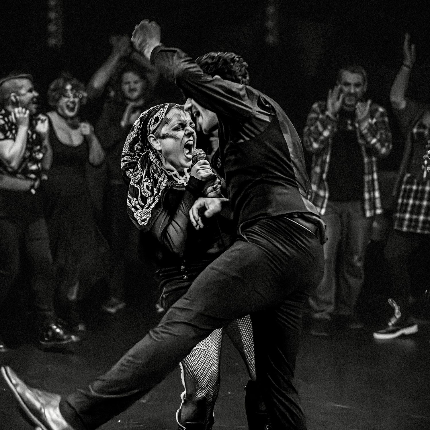 People dance energetically on a black and white dance floor with a crowd watching in the background.