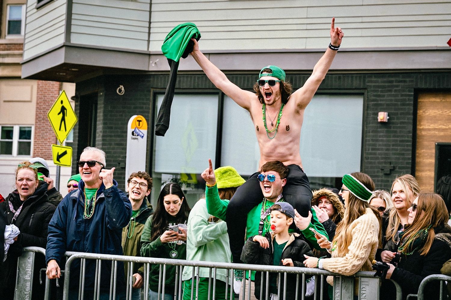 A crowd of excited people in green clothing and accessories celebrate St. Patrick's Day on a city street.