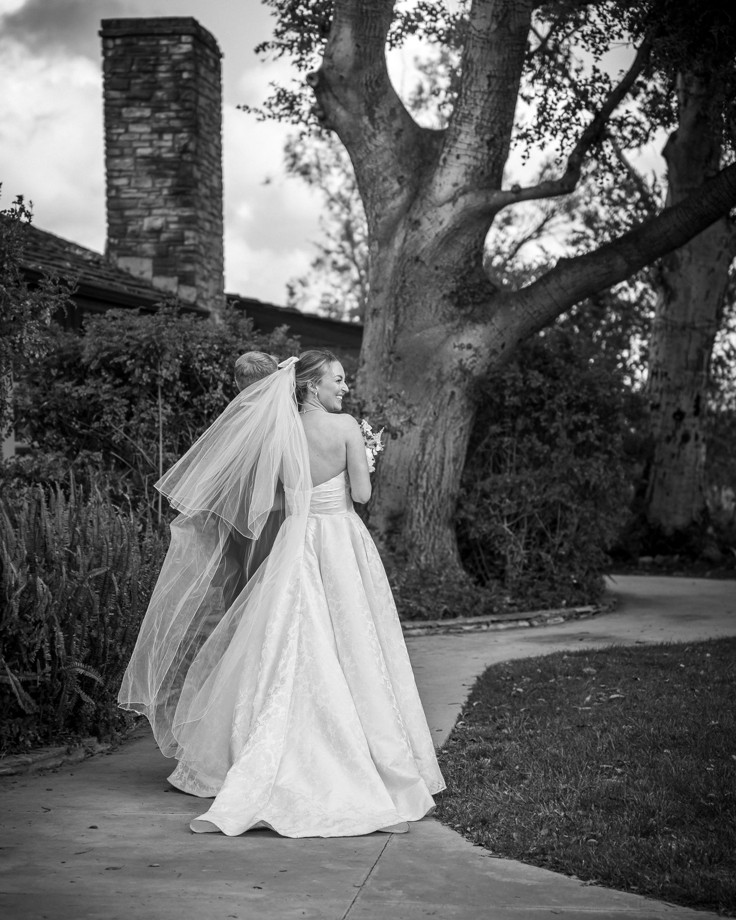A bride in a classic white ball gown and flowing veil stands against a tree beside an old brick chimney in black and white.