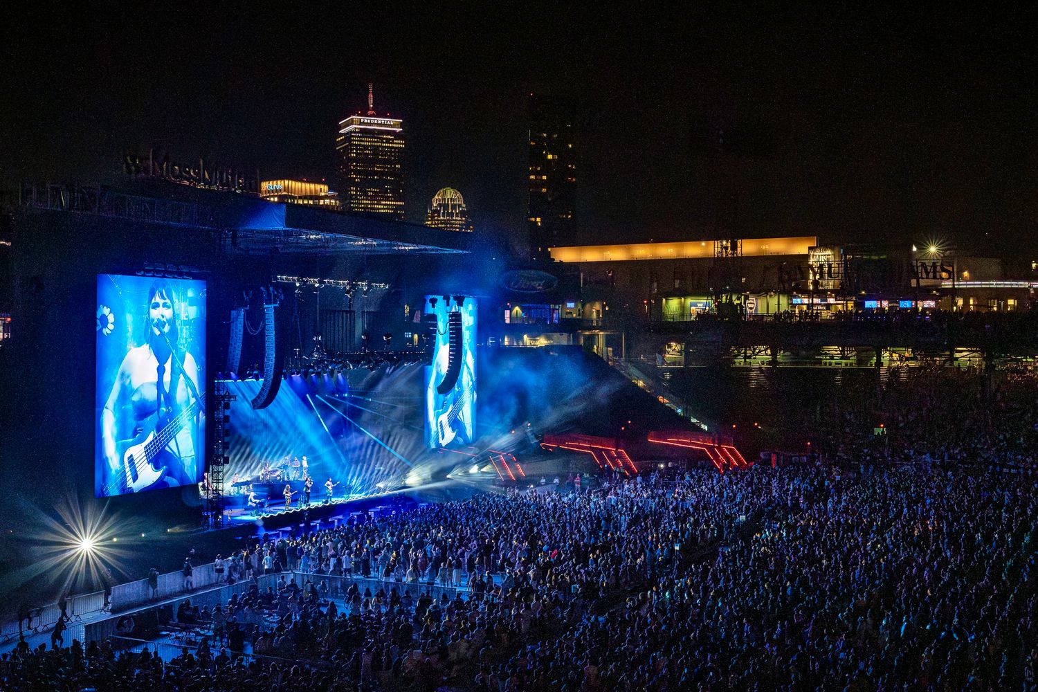 Large outdoor concert stage lit with blue lights performing for massive crowd at night with city skyline visible in background.