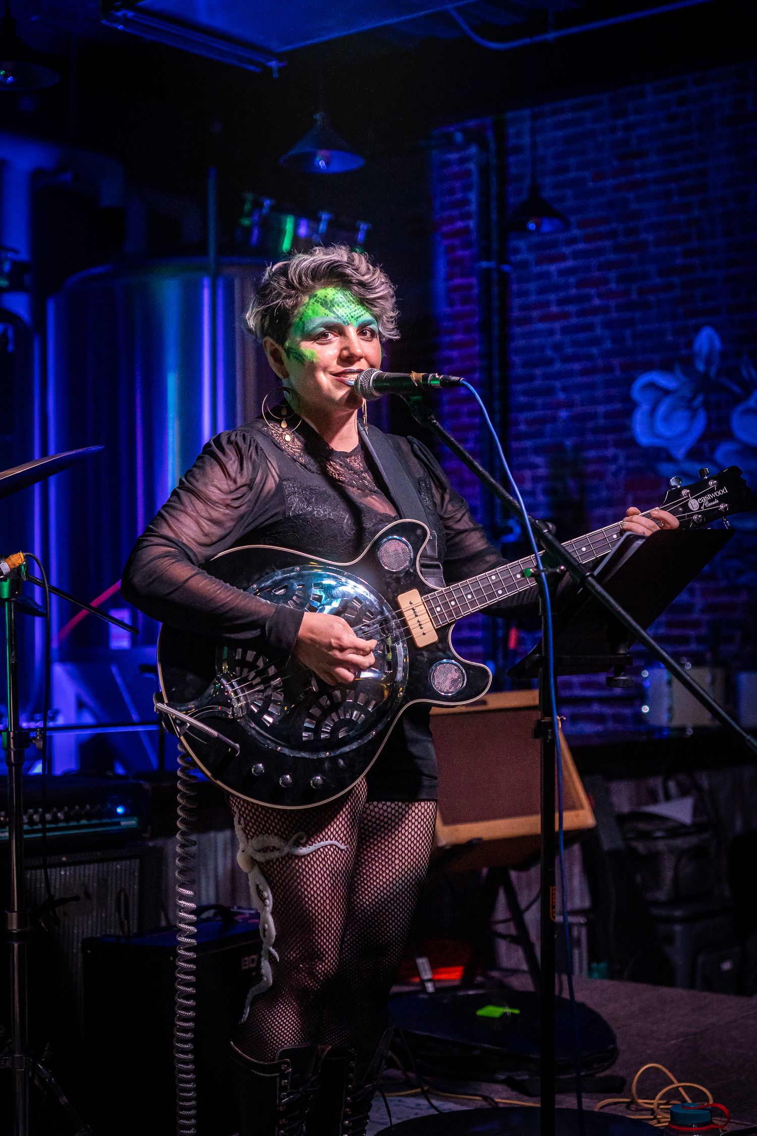 Musician playing resonator guitar performs under blue stage lights in brick-walled venue.