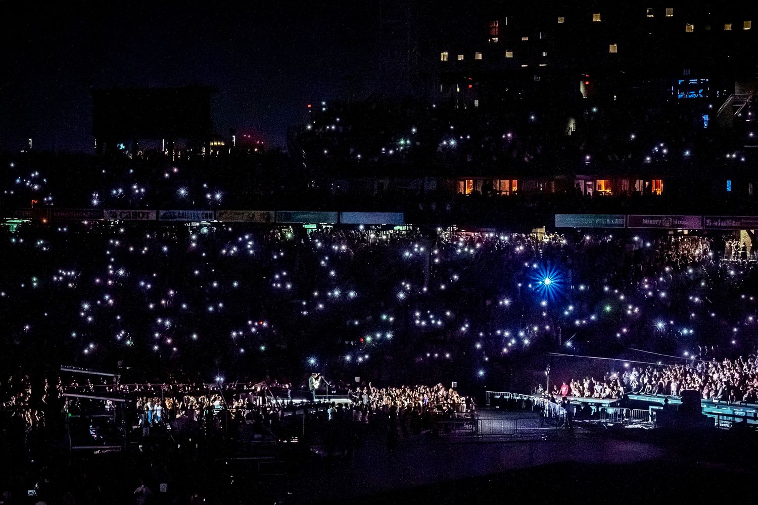 A concert crowd illuminated by blue lights and phone flashlights creates a twinkling sea of lights in a darkened venue.