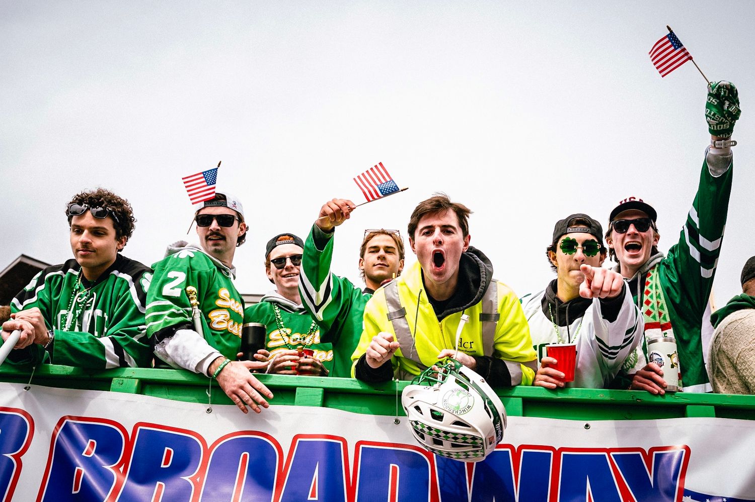Sports fans in green and red jerseys cheer and wave American flags while celebrating at an outdoor event.