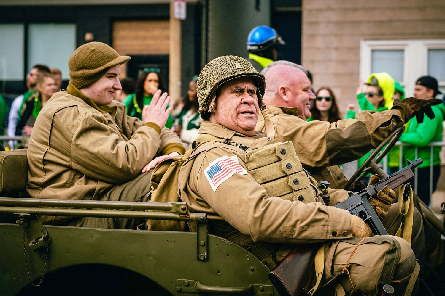 Military veterans in tan uniforms wave to crowds from vintage military vehicles during a parade celebration.