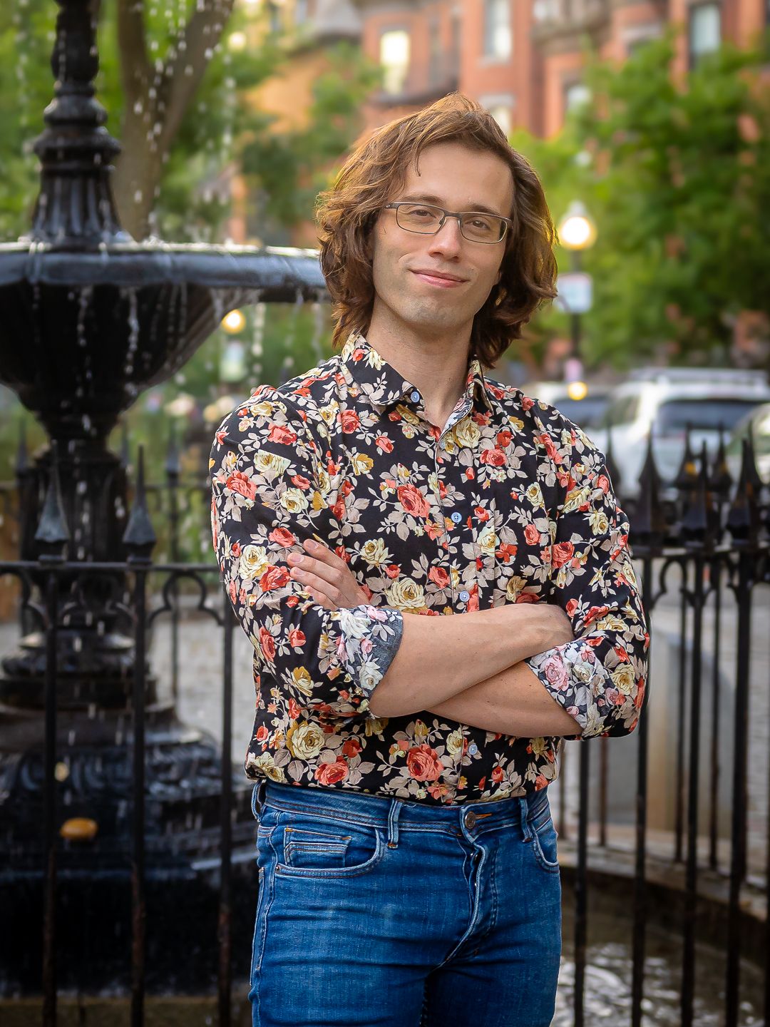 Young man with glasses in floral shirt stands arms crossed by ornate fountain in urban setting.
