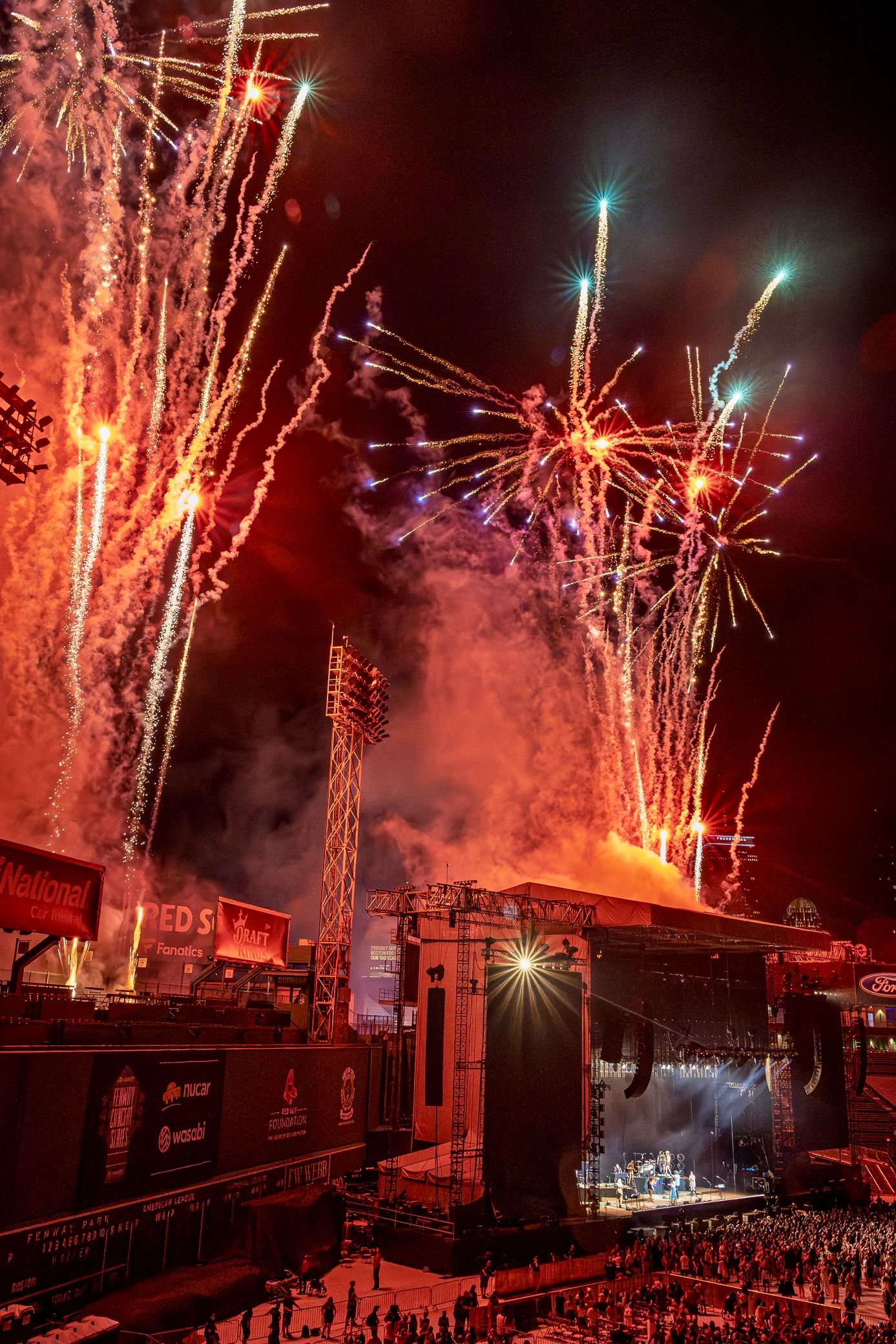 Colorful fireworks explode over an outdoor concert venue with stadium lights illuminating the stage below during a nighttime show.
