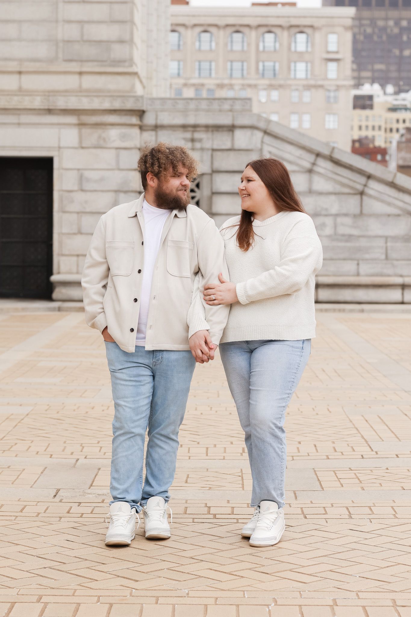 A couple in light-colored clothing holding hands while standing on brick pavement near urban architecture.