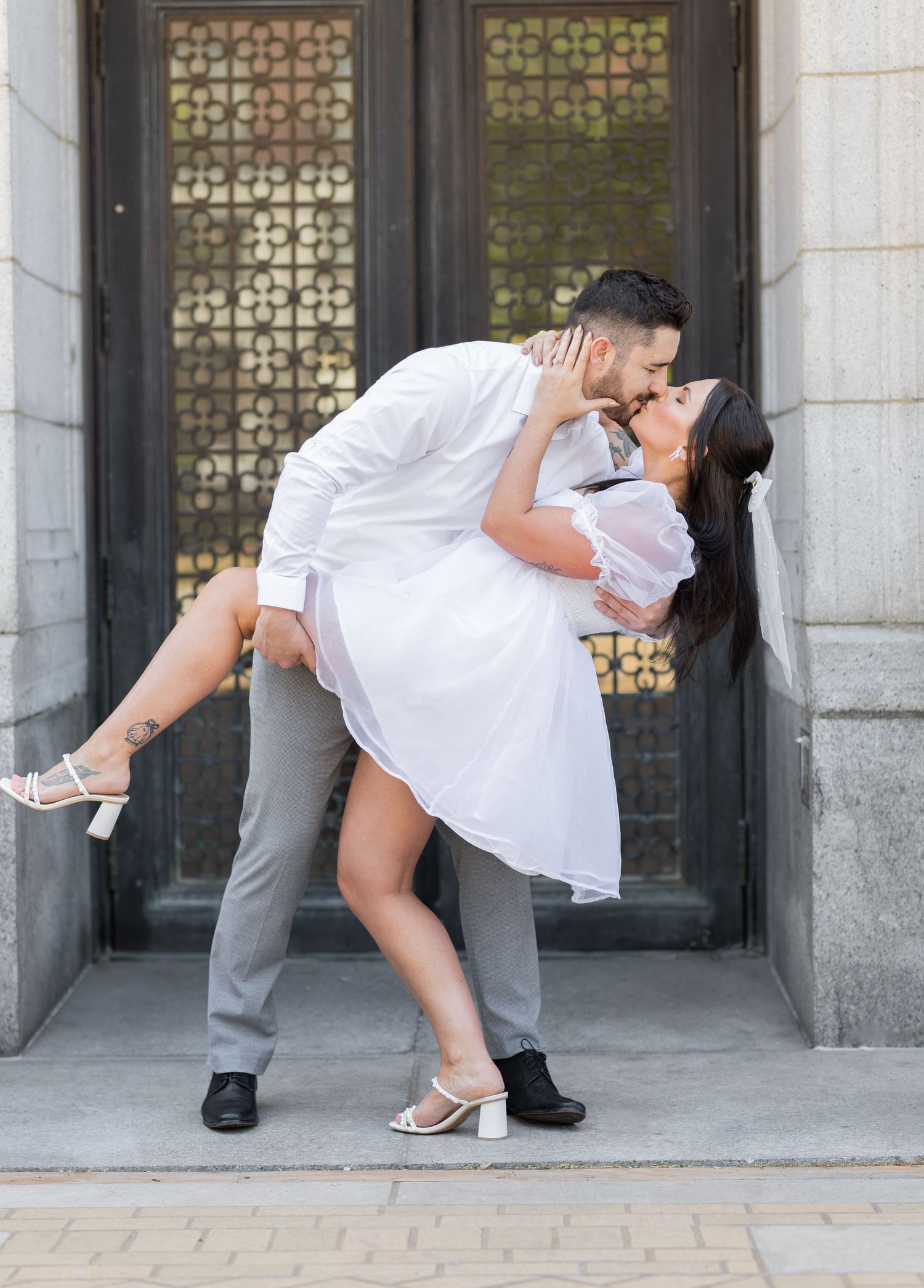 A romantic dip pose between a couple in matching white outfits against an ornate doorway.