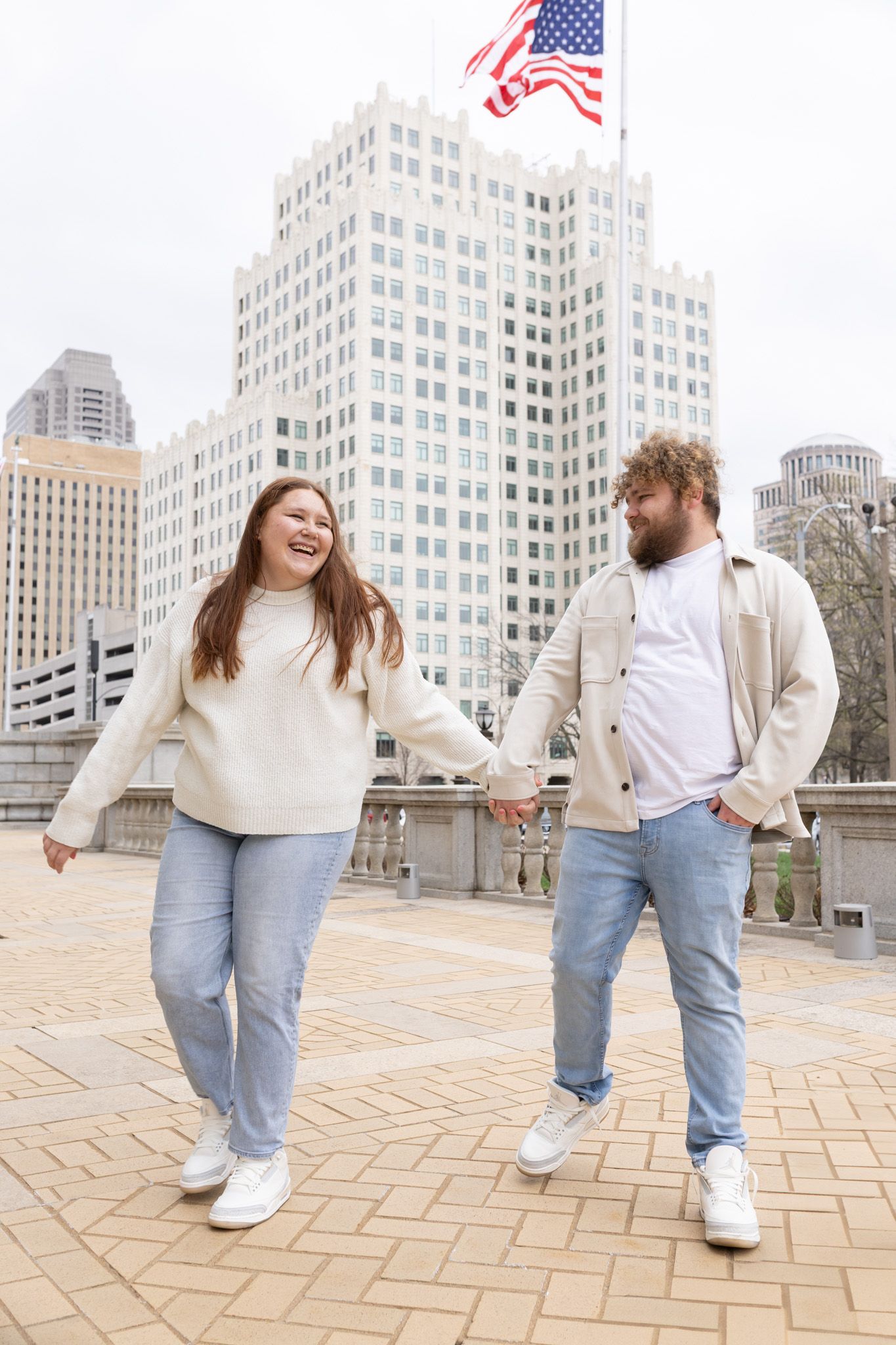 A couple in coordinating casual outfits laughing together in front of a city building with an American flag.