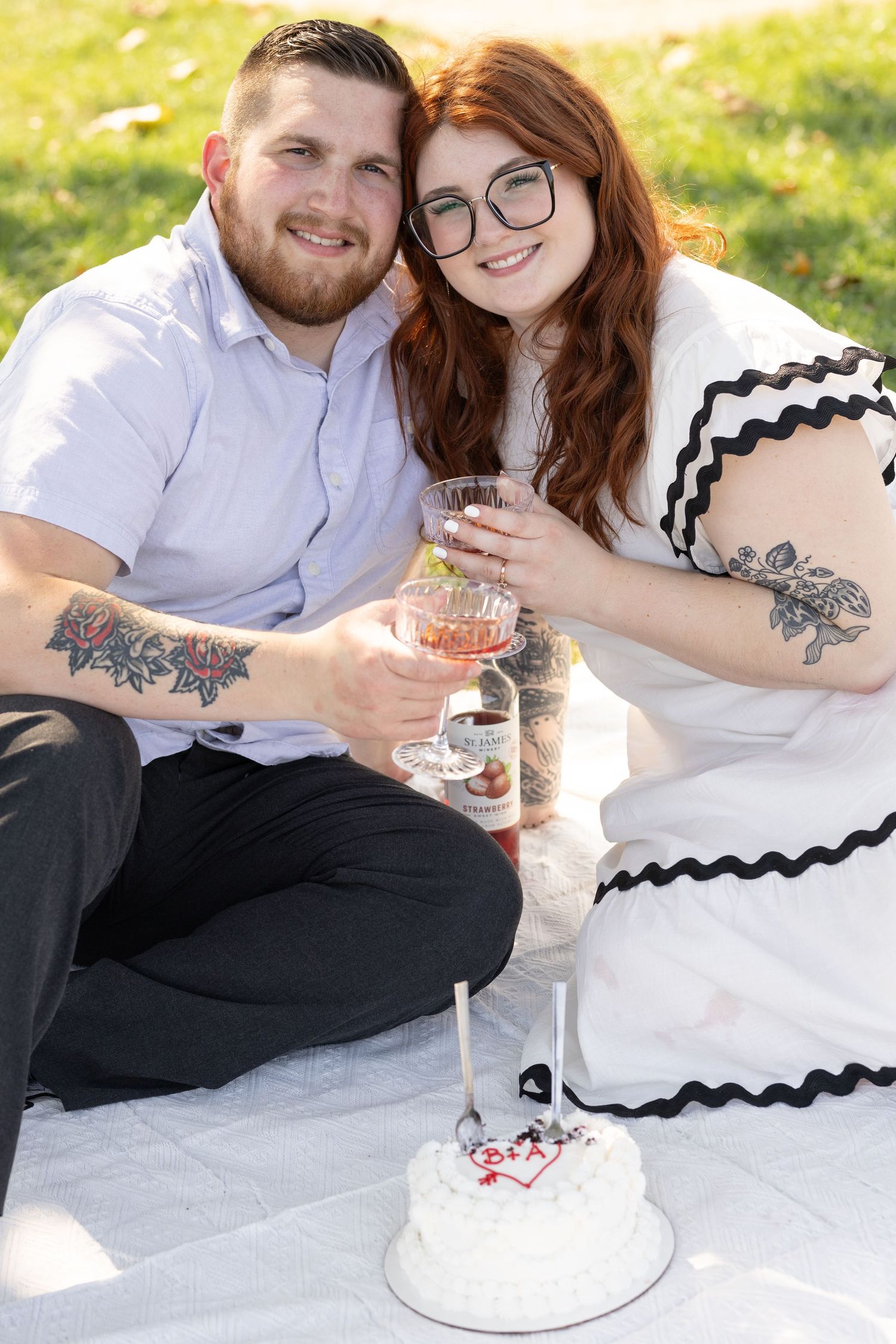 Couple toasting with drinks at outdoor picnic with small white cake.