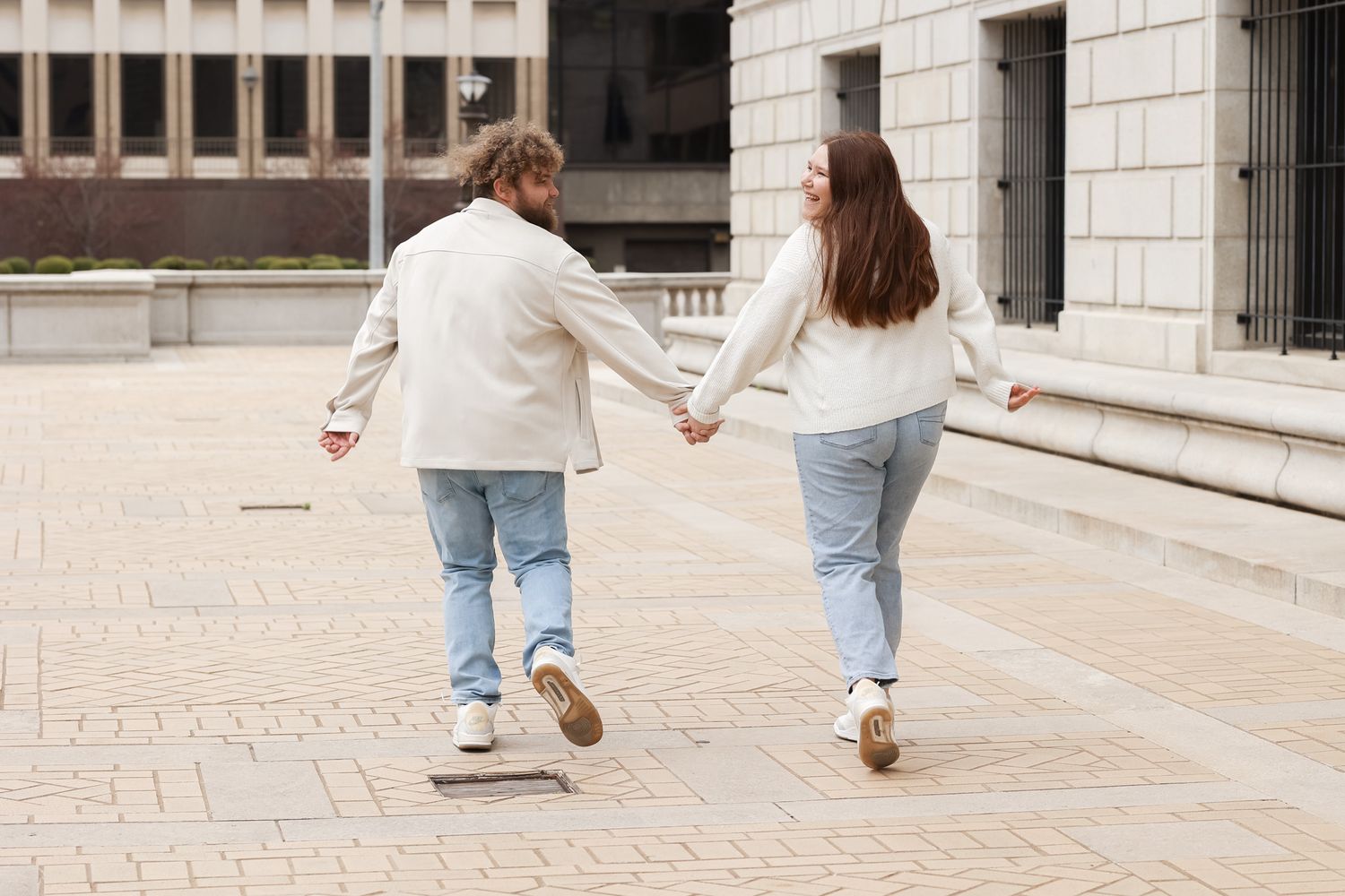 A couple holds hands while walking away from camera on a brick plaza in matching casual outfits.