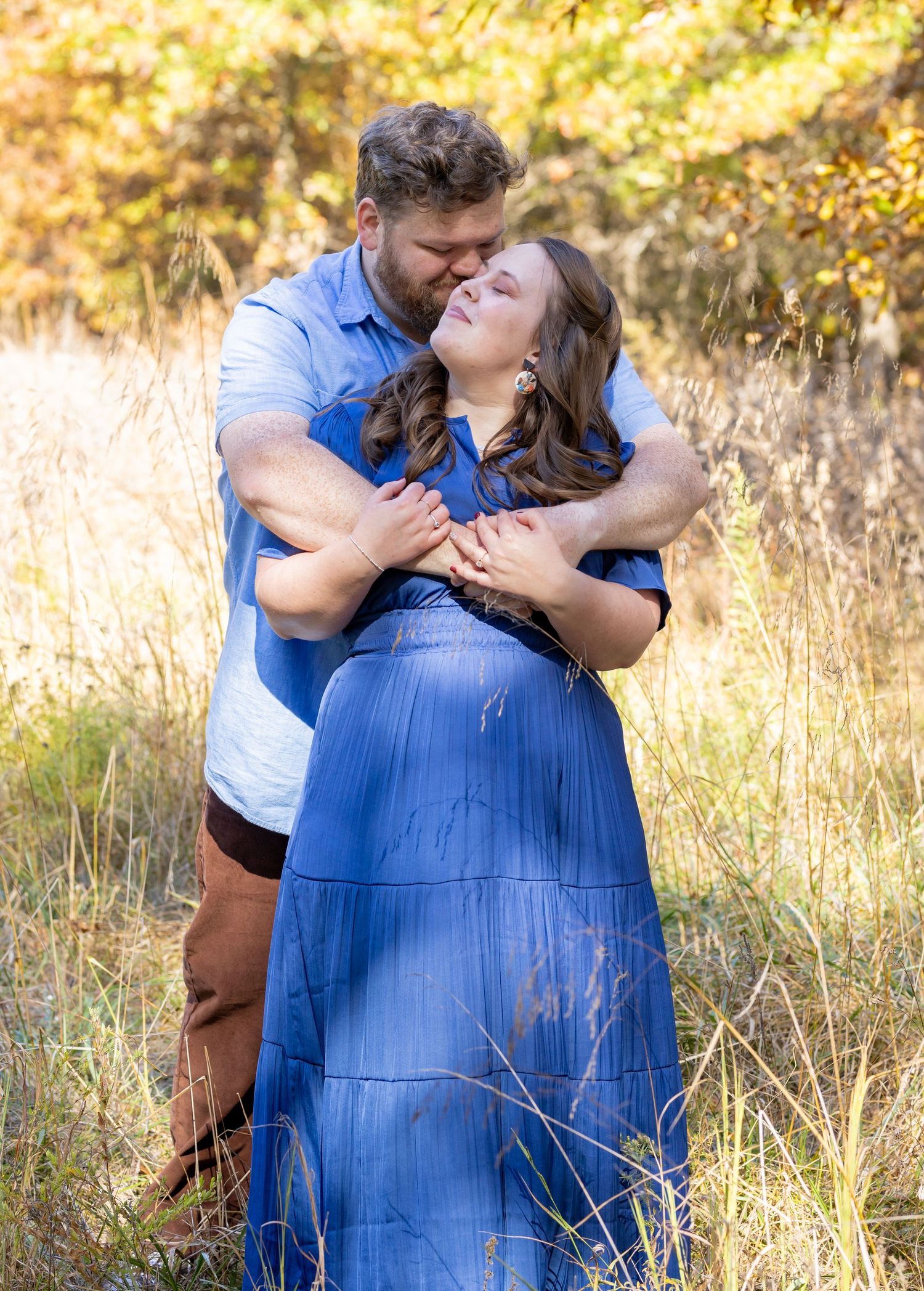 Romantic couple embracing in golden field wearing blue dress and brown pants.
