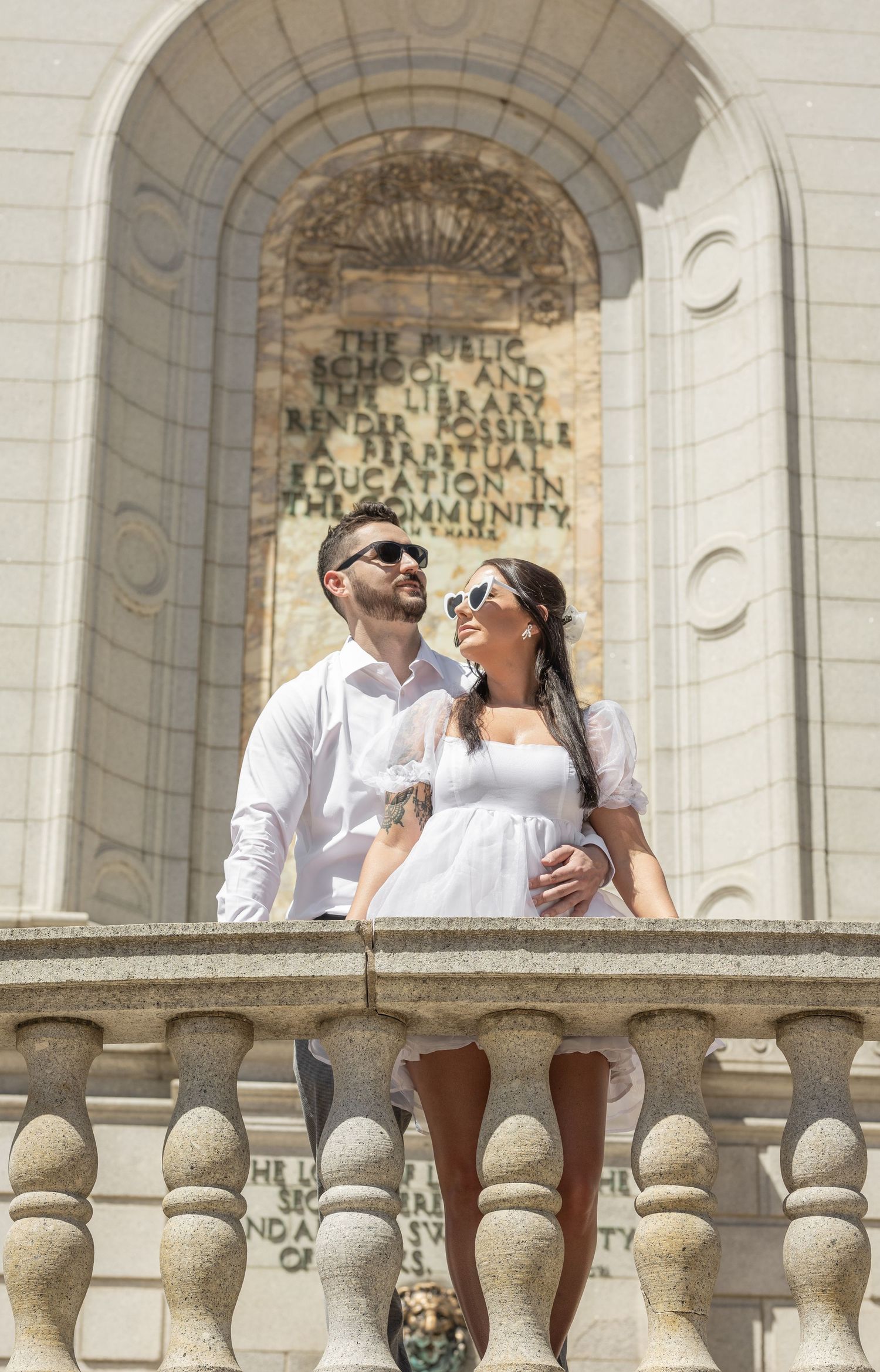 A couple in white outfits sharing a romantic moment on a classical stone balcony.