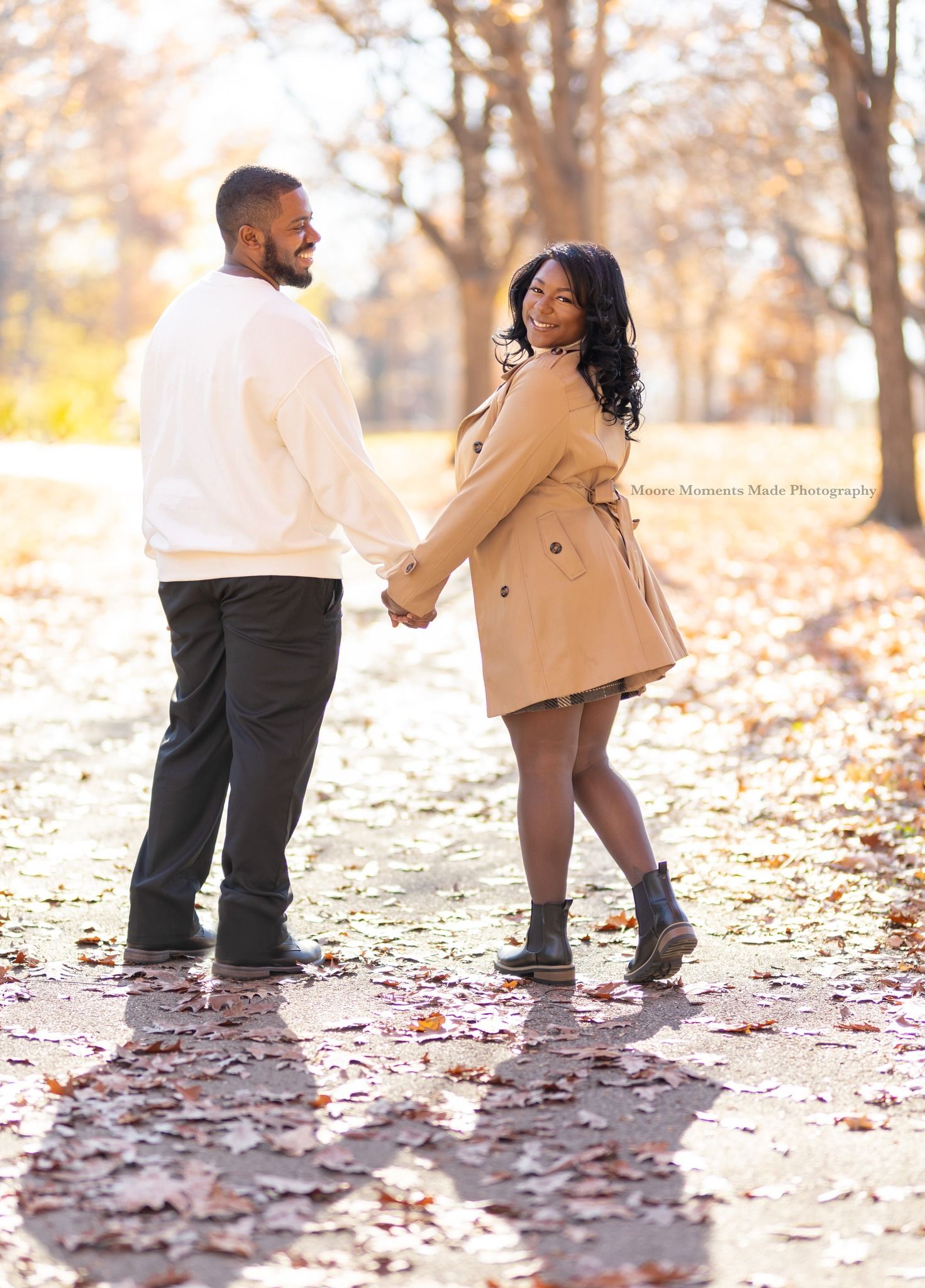 A romantic autumn engagement photo of a couple walking through fallen leaves in a park setting.