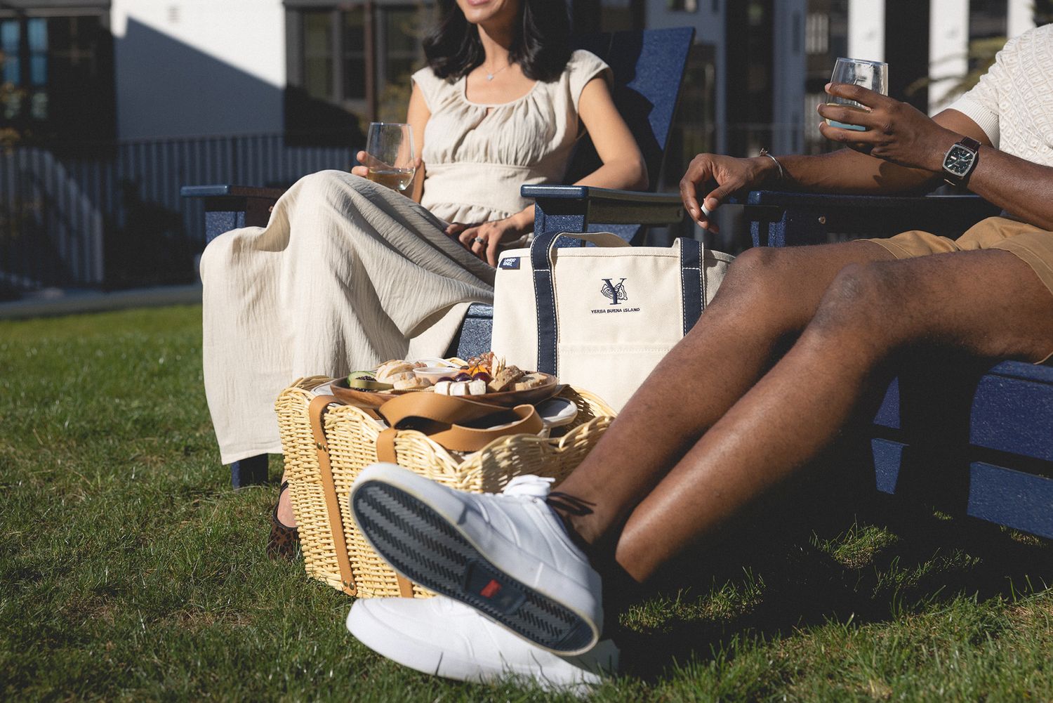 Two women enjoying a sunny outdoor picnic on grass chairs with a wicker basket and tote bag nearby.