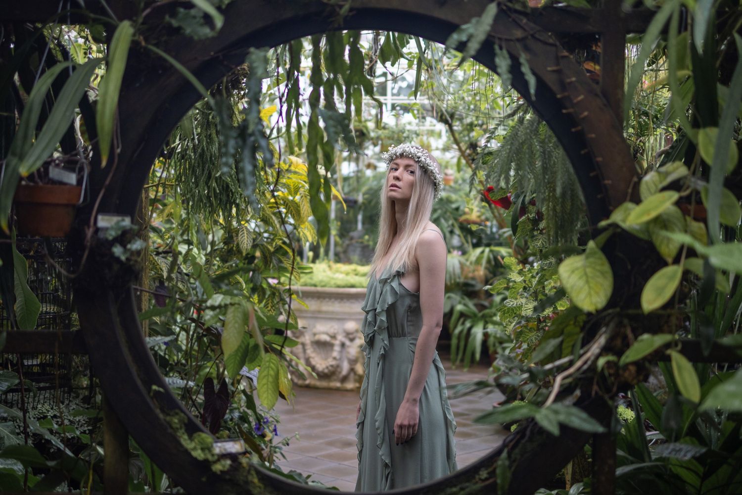 Blonde woman in green dress standing inside a circular garden archway surrounded by lush tropical plants.