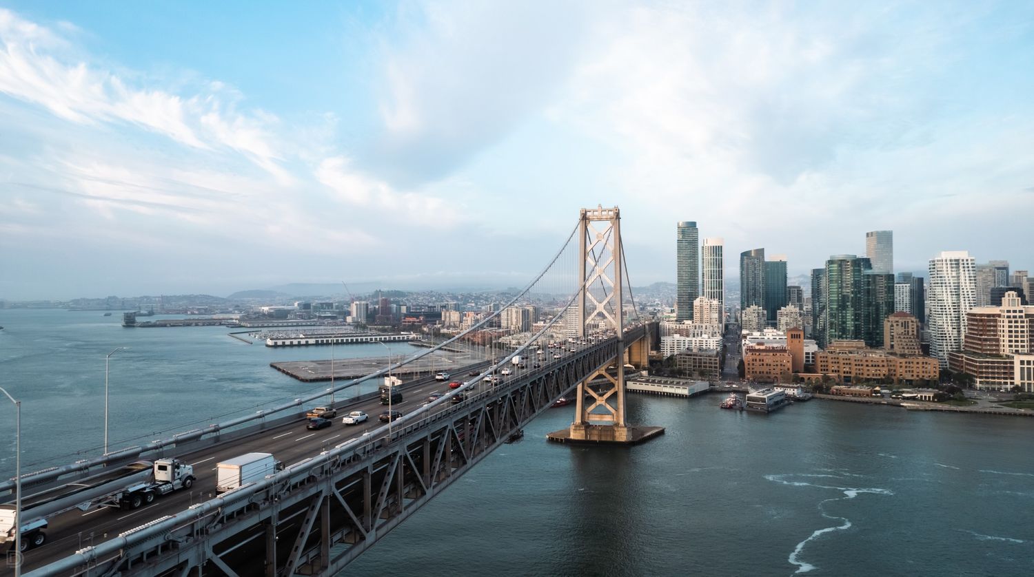 Aerial daytime view of San Francisco Bay Bridge with city skyline and waterfront under partly cloudy blue sky.