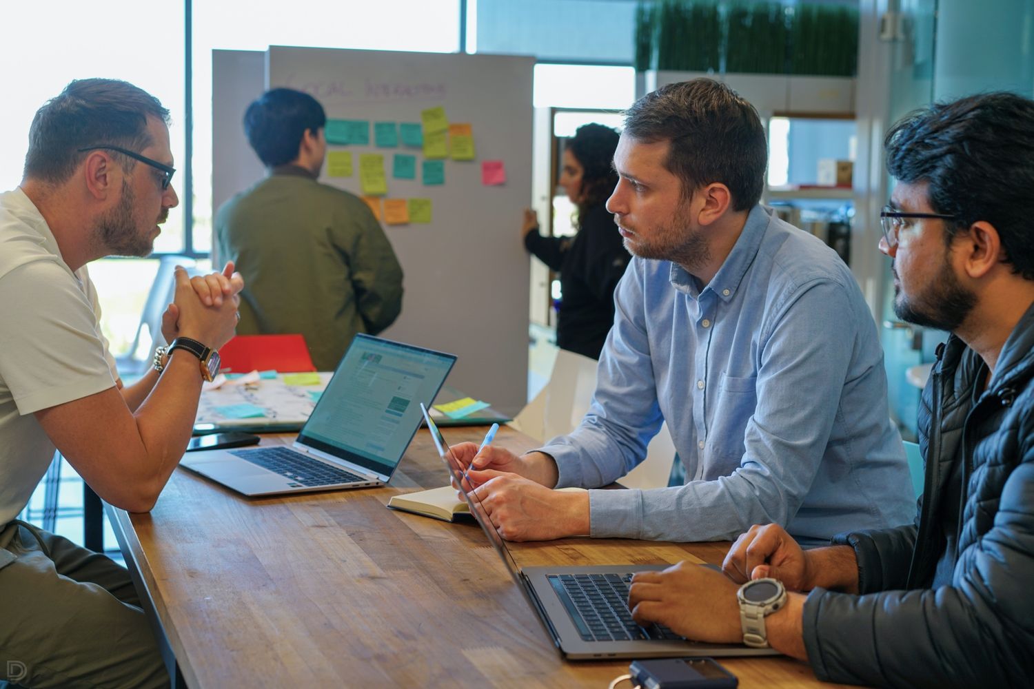 Three professionals collaborating around laptops at a wooden table in a modern office workspace.