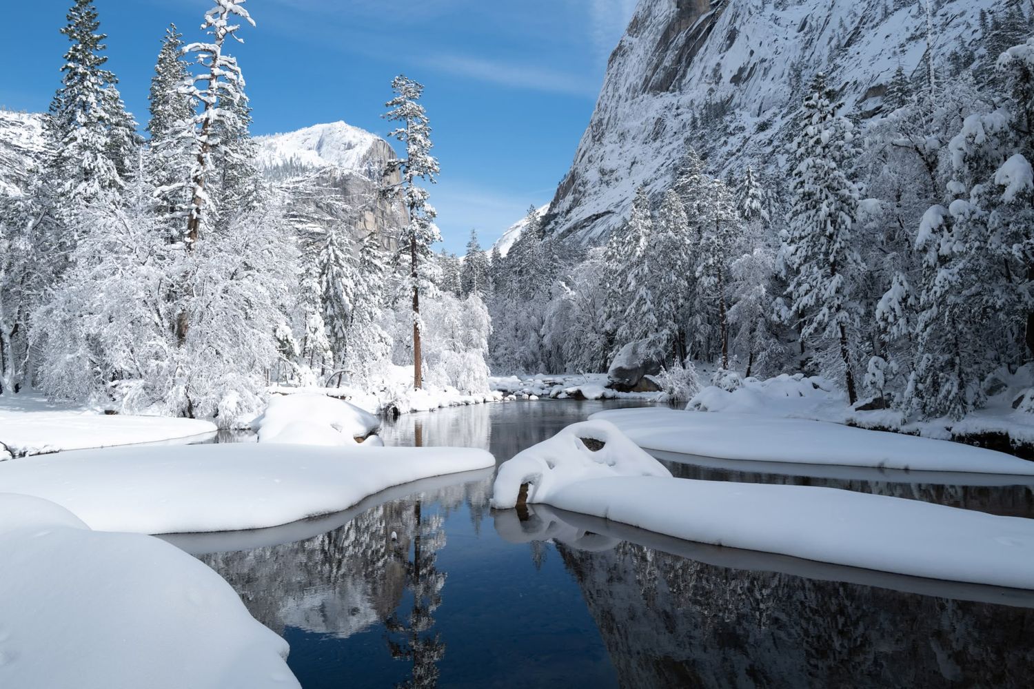 Serene winter scene in Yosemite with snow-covered trees reflected in a calm river under blue skies.