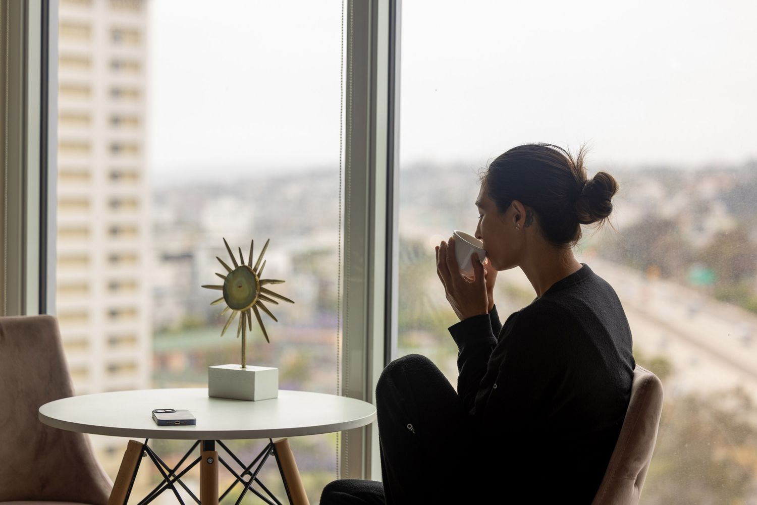 Woman in black outfit sits contemplatively by large window with city skyline view and decorative sun sculpture on table.