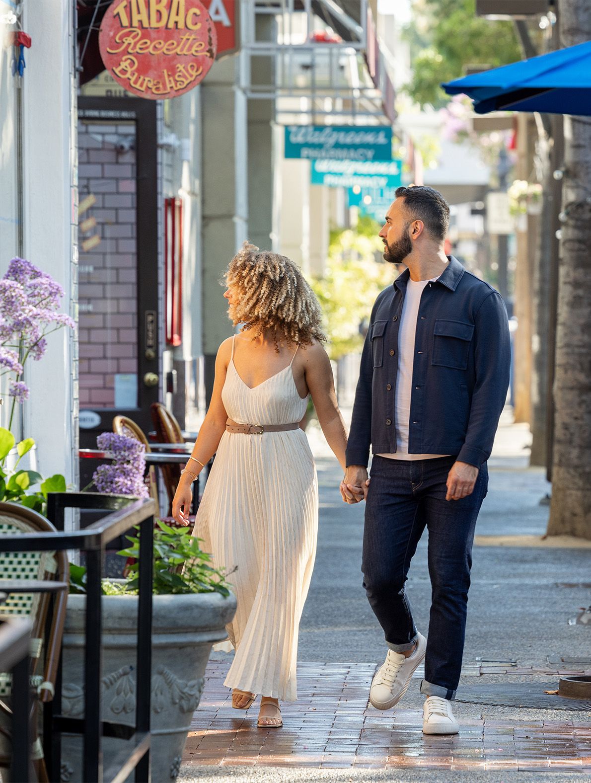 Couple holding hands walking on a sunny urban sidewalk, woman in white dress, man in navy jacket and jeans.