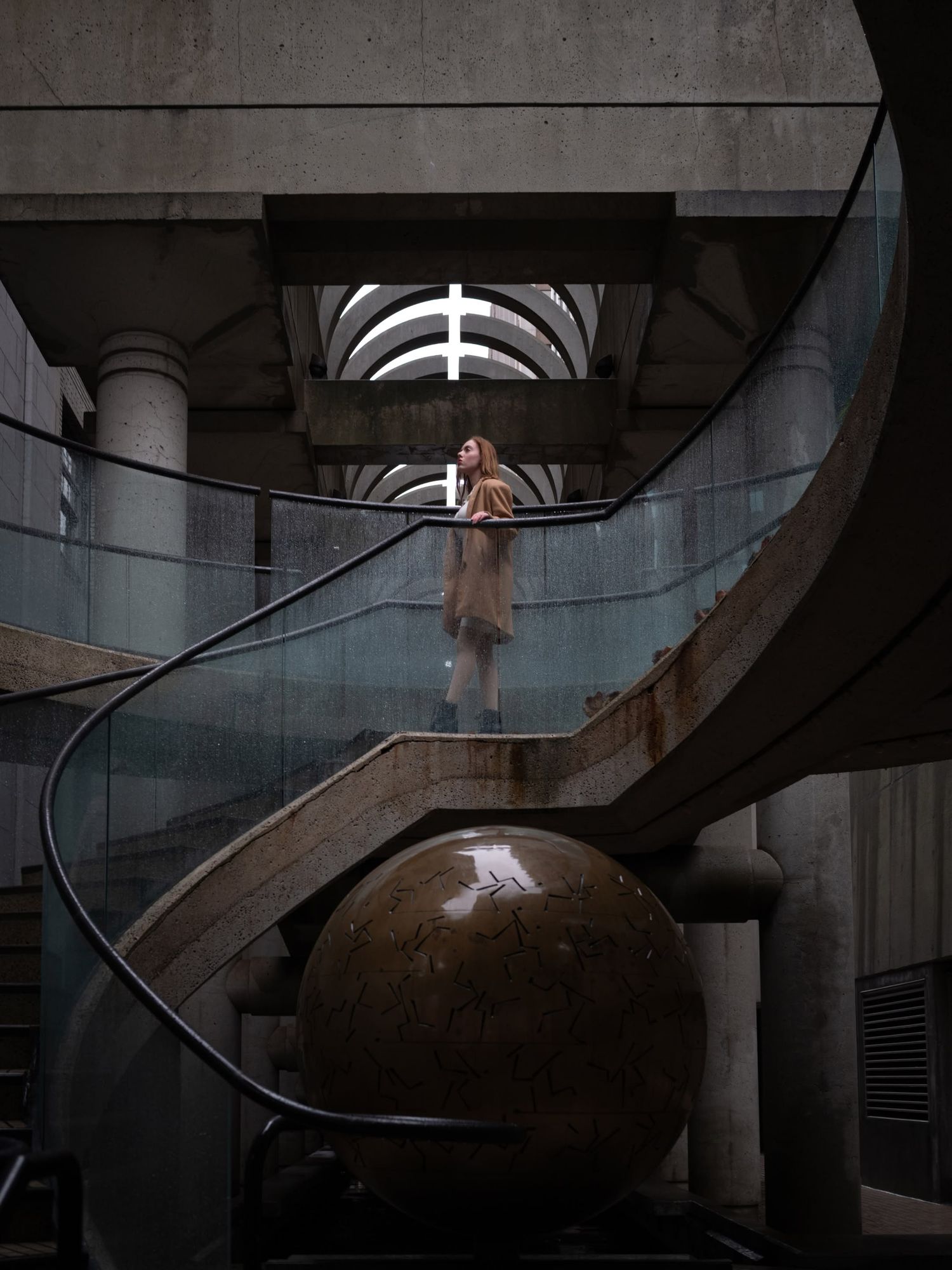 Woman standing on a spiral staircase above a large bronze sphere in a dramatic brutalist building.
