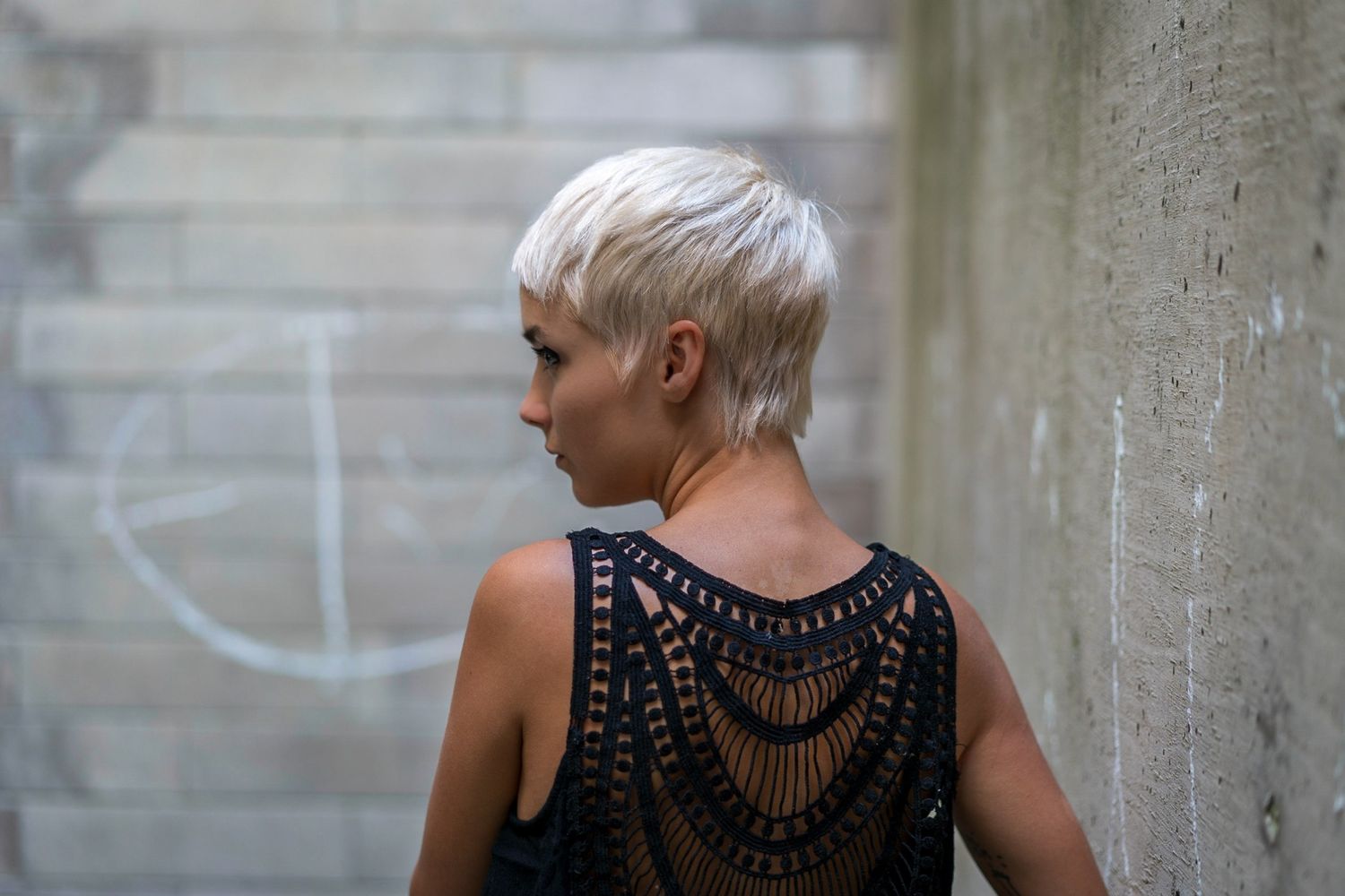Woman with platinum pixie cut wearing black crochet top viewed from behind against concrete wall.