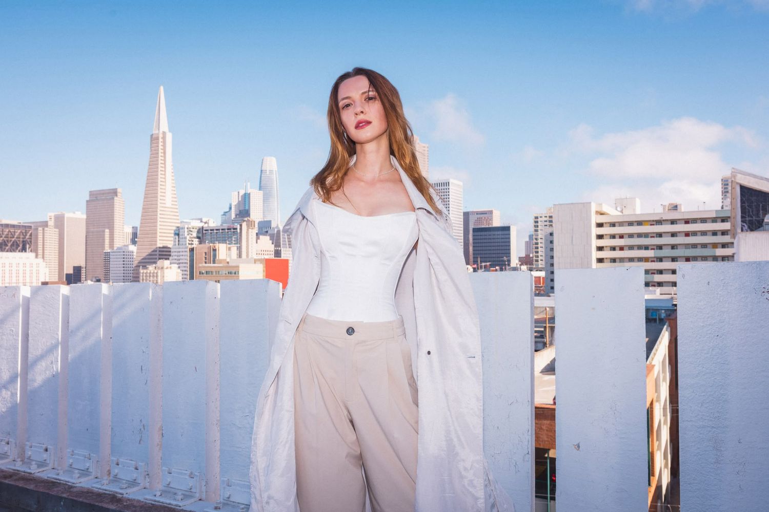 Young woman in white corset top and beige trench coat poses on rooftop with San Francisco skyline behind her.