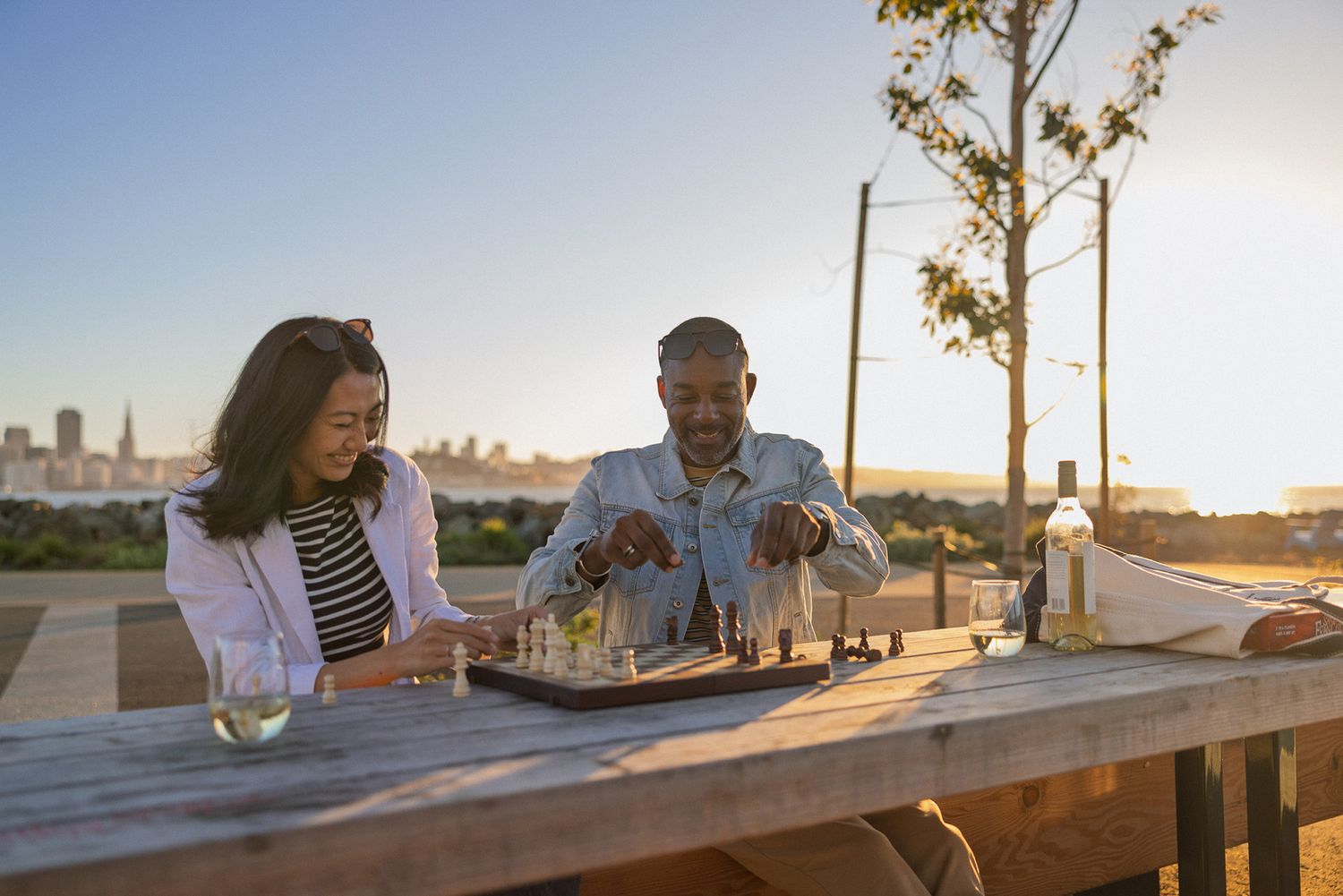 Couple playing chess at outdoor wooden table during golden sunset with city skyline in the distance.