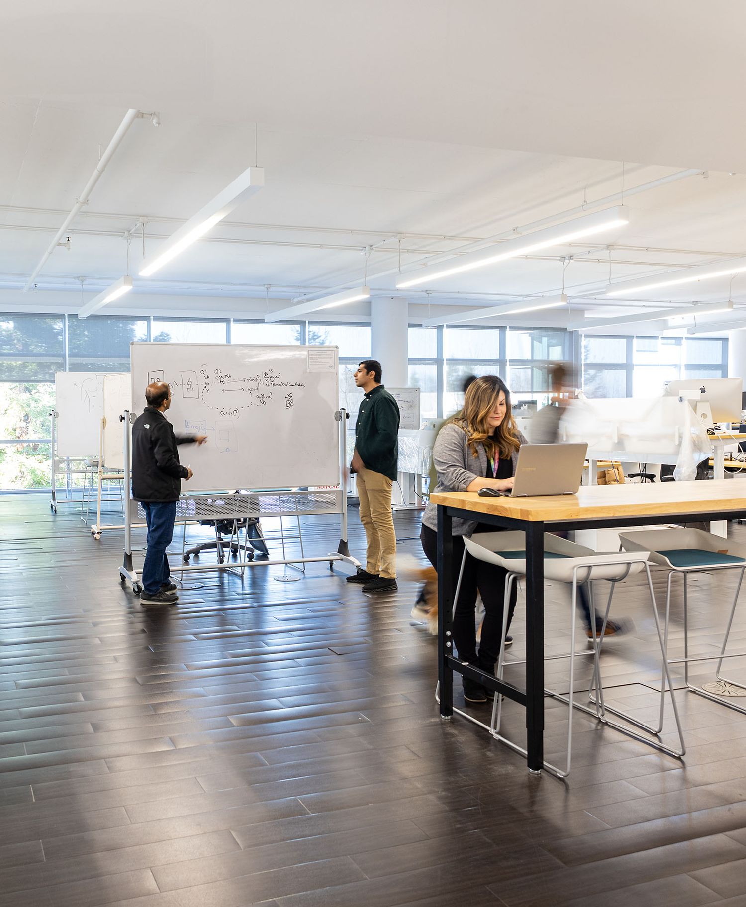 Professionals collaborate in a modern open office space with whiteboards and standing desks under bright lighting.