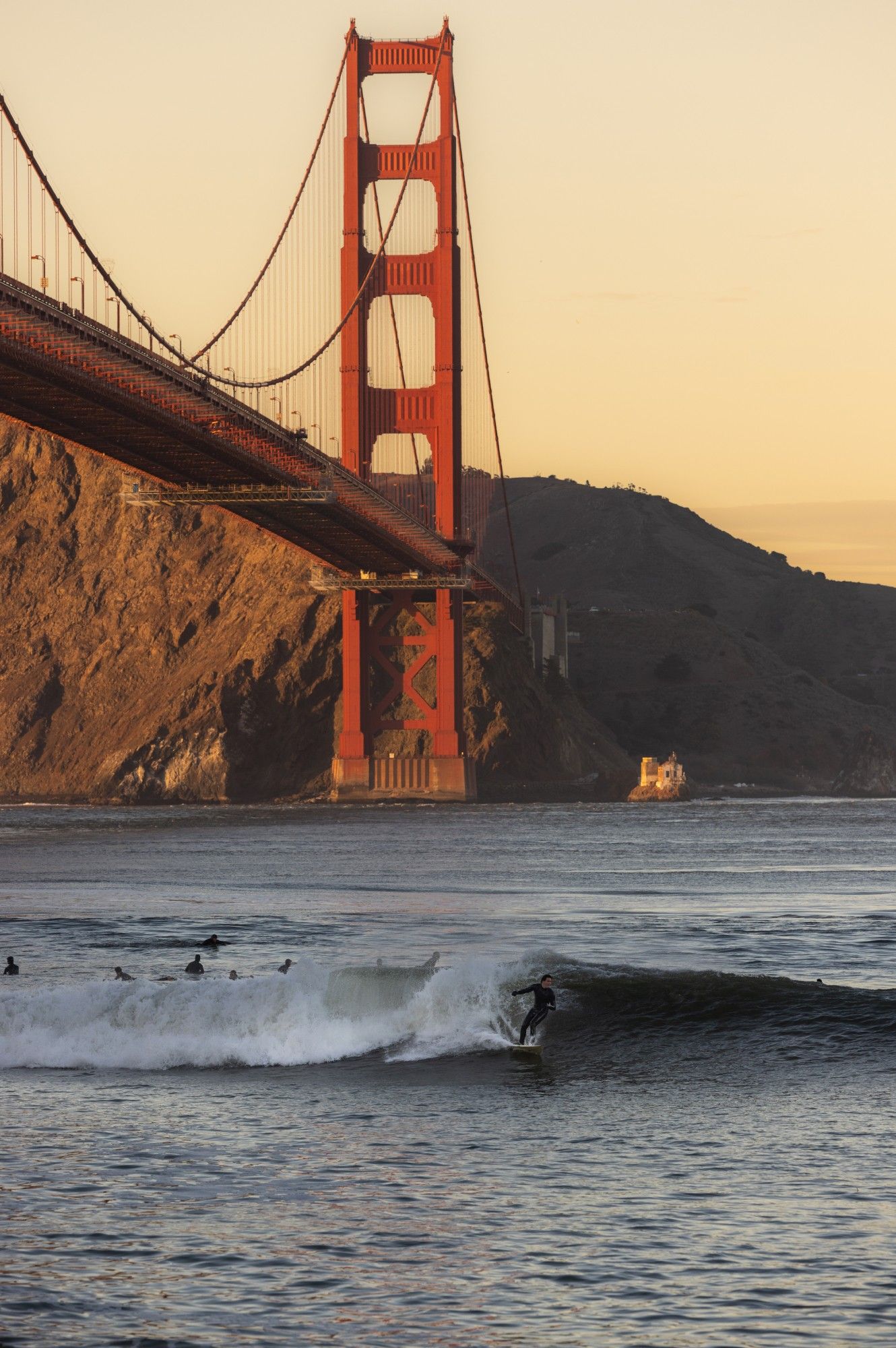 Surfer riding waves near the iconic Golden Gate Bridge in San Francisco at golden hour with hills in background.