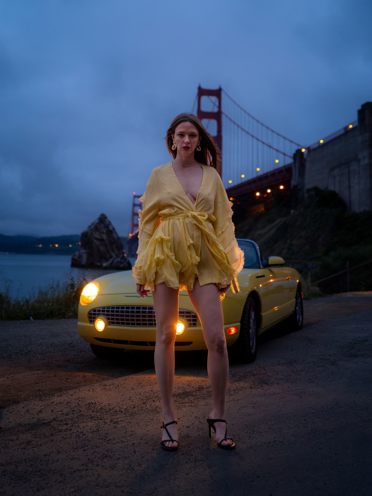 Woman in yellow dress poses before a yellow convertible with Golden Gate Bridge lit up at dusk.