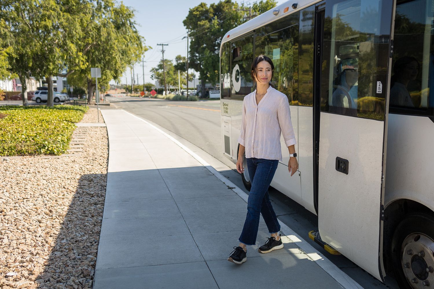 Woman stepping off a modern white bus onto a sidewalk in a sunny suburban area with trees and landscaping.