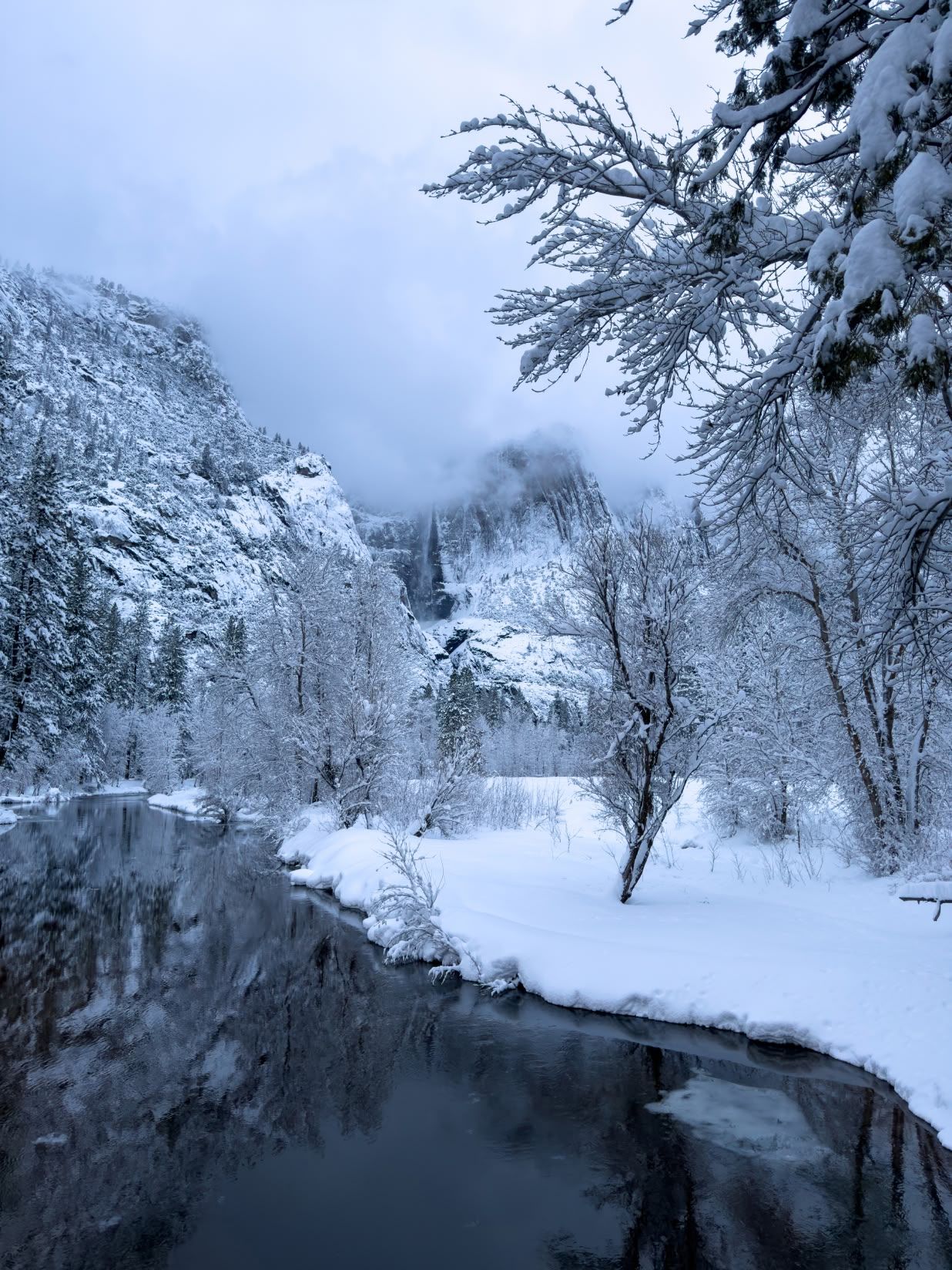 Misty winter landscape along Merced River in Yosemite with snow-covered trees and cloudy mountain views.