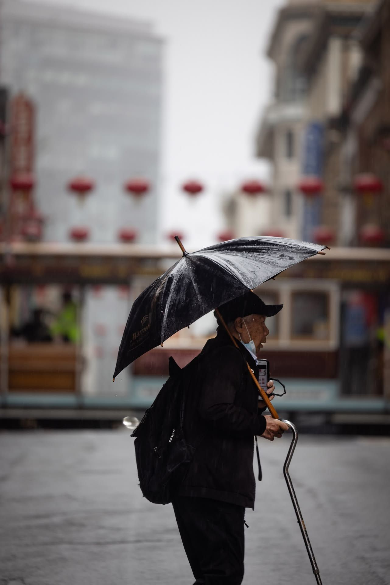 Elderly person walking in the rain with a black umbrella near red lanterns in Chinatown.