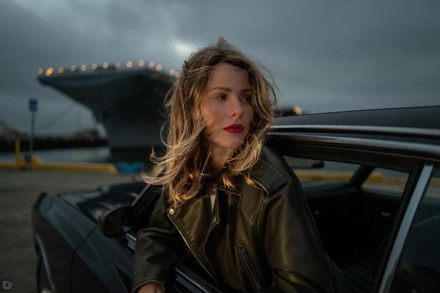 Woman in leather jacket exiting a dark car with a large naval ship in the cloudy background.