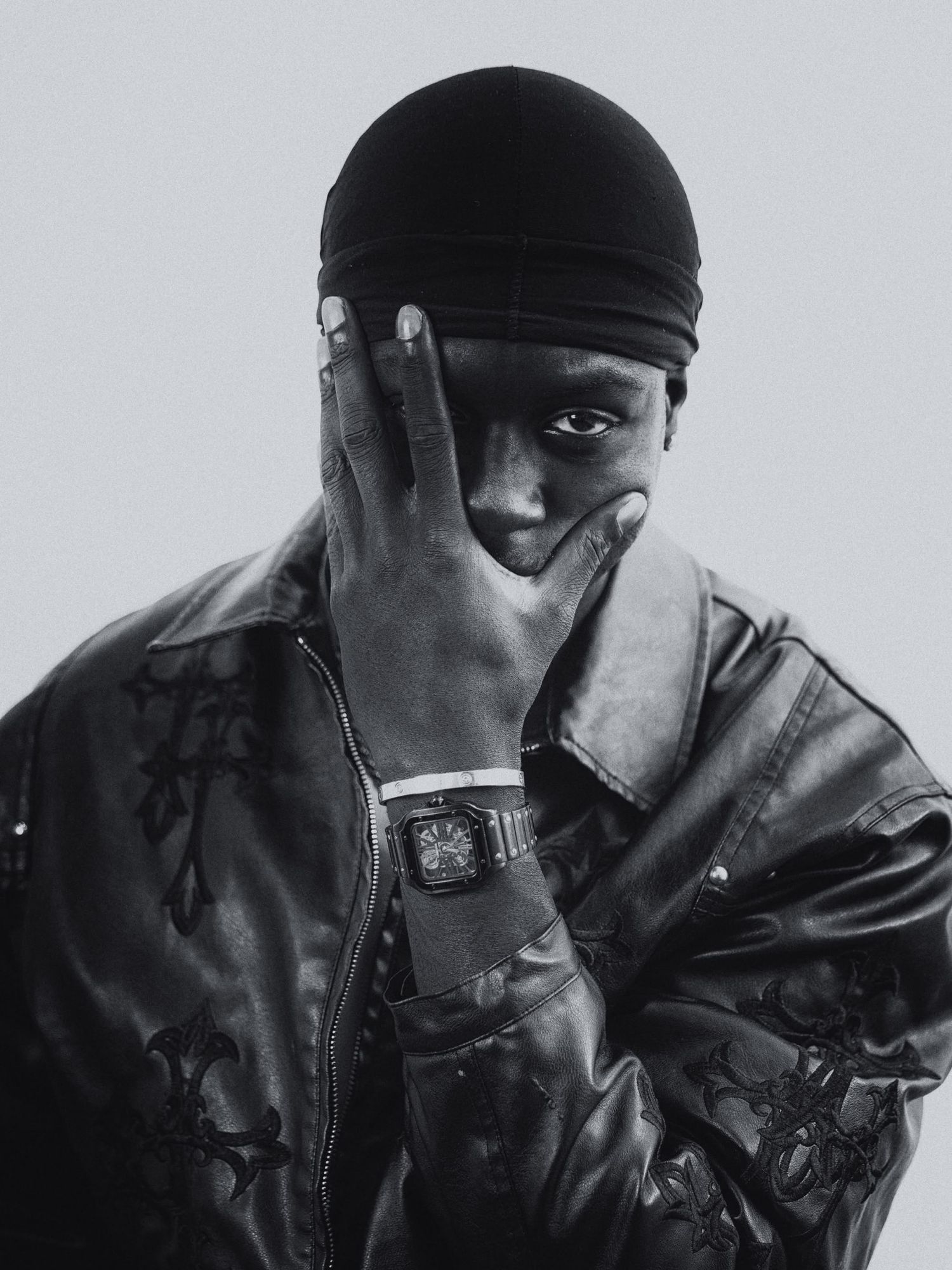 Black and white portrait of a man in leather jacket and durag covering his face with his hand.