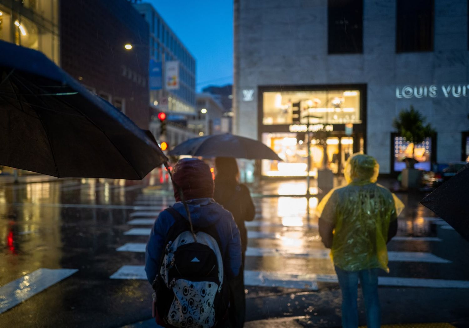 Two people with umbrellas waiting at a rainy crosswalk near a Louis Vuitton store at dusk in the city.